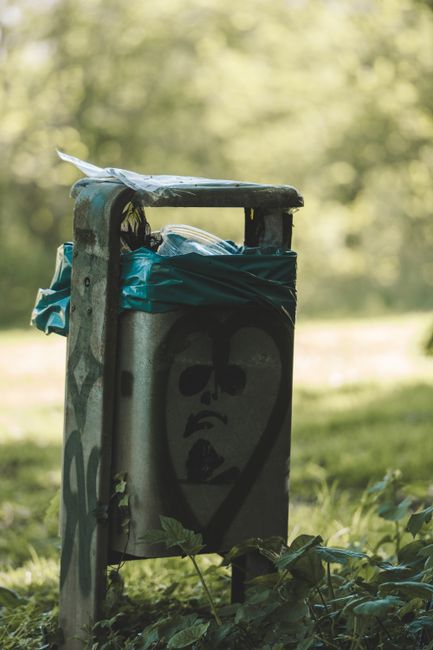 A weathered grey metal mailbox with a black hinged lid, mounted on a slanted post that is partially embedded in the ground, situated in a forested area with dense, green foliage and tall trees in the 