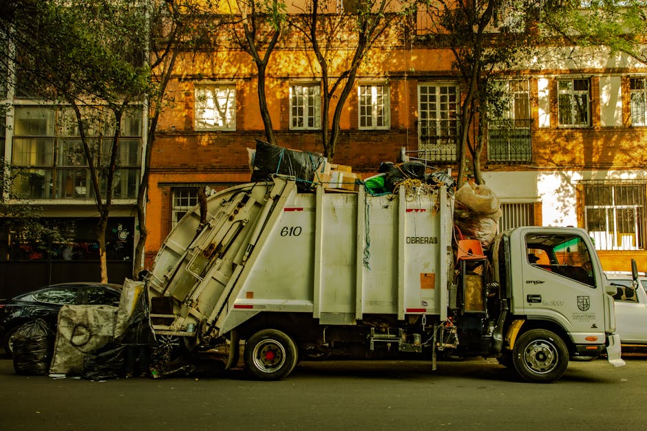 A scene of waste accumulation alongside a dirt road with a small pile of rubbish consisting of dry, brown leaves, discarded plastic containers, and paper debris in the foreground. Behind the rubbish p